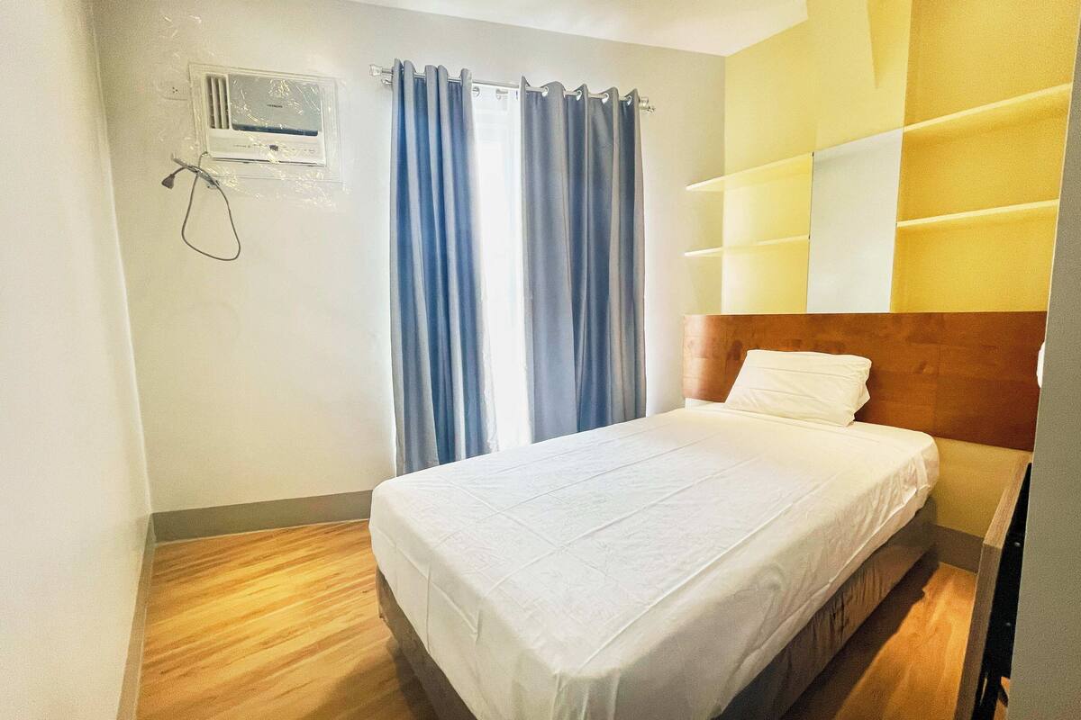 A hotel room with a bed, a window with curtains, a wall-mounted air conditioning unit, and a wooden headboard.