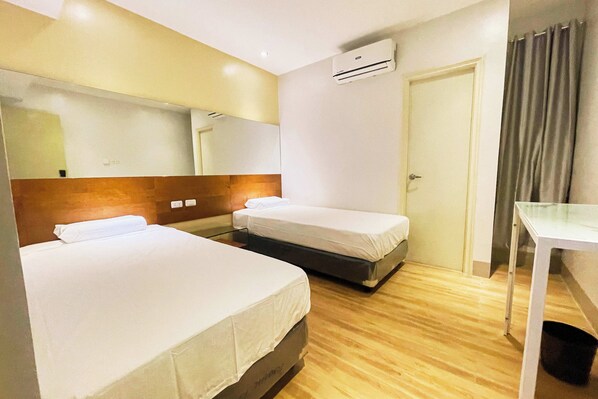 Una habitación de hotel con dos camas individuales, cabecera de madera, escritorio blanco, espejo y unidad de aire acondicionado.