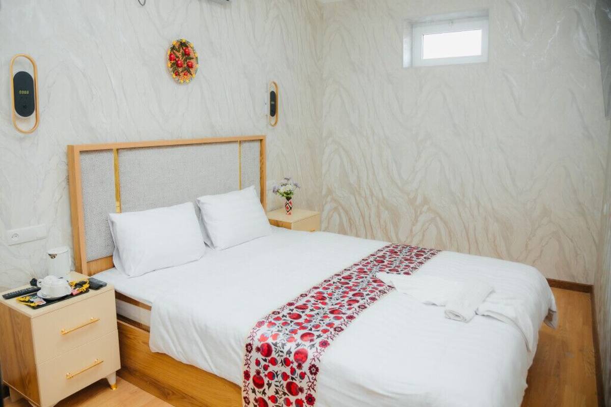 A neatly made bed with white bedding and a red and white floral blanket, a bedside table with a lamp, and a small window with a view of the outside.