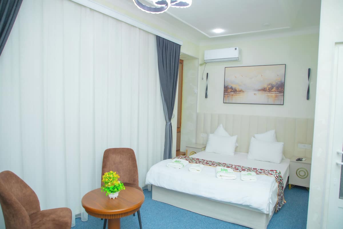 A hotel room with a bed, a small table with a plant, two chairs, a painting on the wall, and an air conditioning unit.