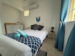 A bedroom with a bed, a bedside table, a lamp, a window with curtains, a wall-mounted air conditioner, and a small plant. - Martello Duplex - 1 Min Walk to La Preneuse Blue Sea (La Preneuse)