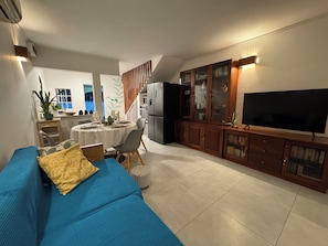 A modern living room with a blue sofa, a dining area with a round table, and a kitchen area with a refrigerator and a TV. - Martello Duplex - 1 Min Walk to La Preneuse Blue Sea (La Preneuse)