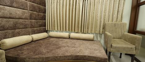 A corner seating area with a brown sofa and a beige armchair, a window with beige curtains, and a wooden floor.