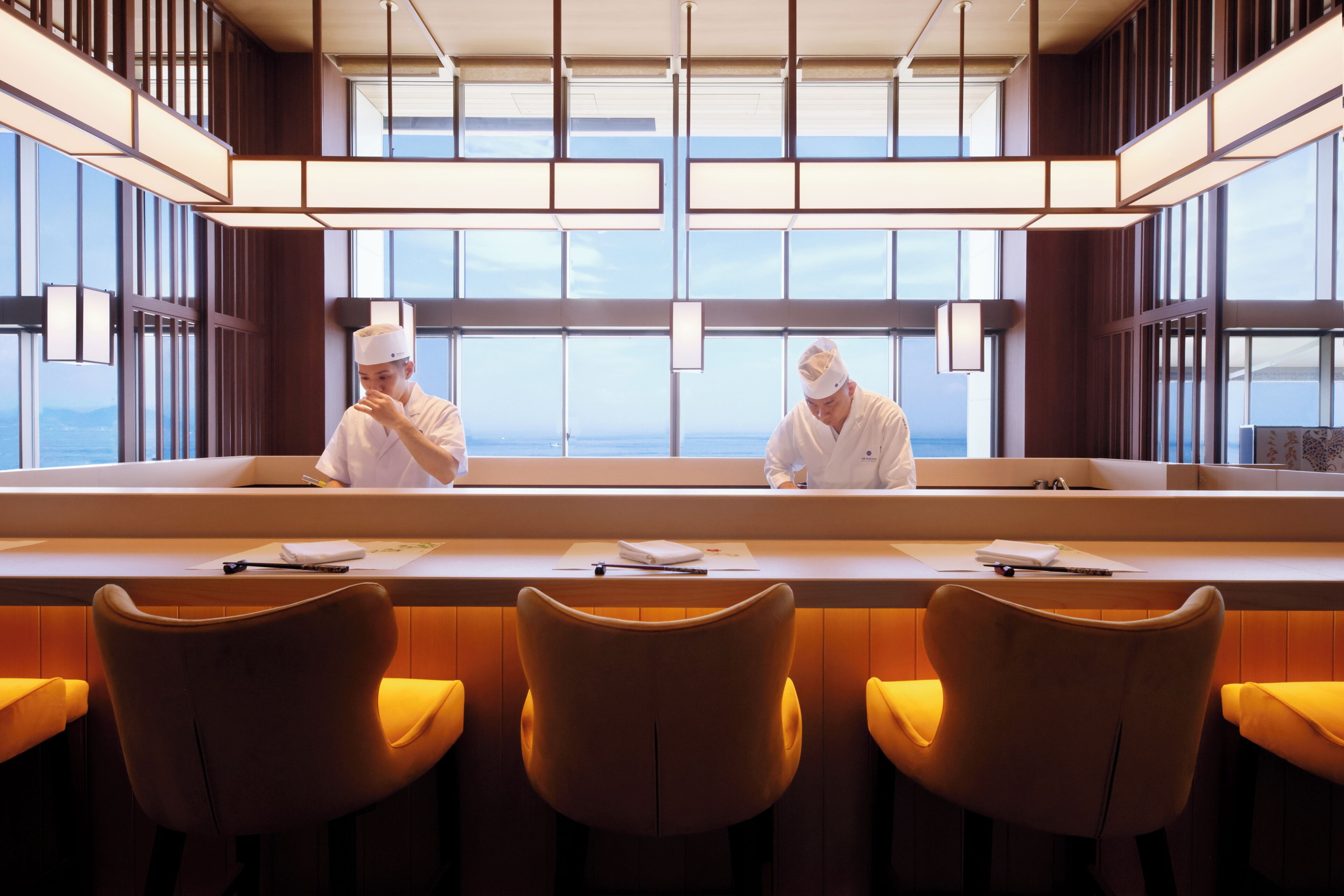 A modern restaurant with a long counter, two chefs, and three bar-style seating areas.