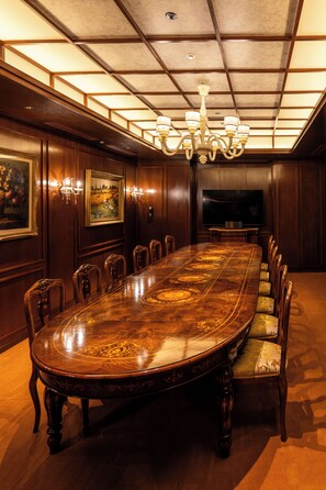 Une salle de conférence avec une longue table en bois, des chaises, un lustre, un tableau et un téléviseur.
