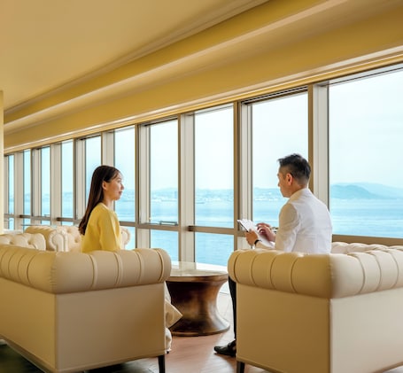 A room with two sofas facing each other, a coffee table, and large windows with a view of the sea and distant land.