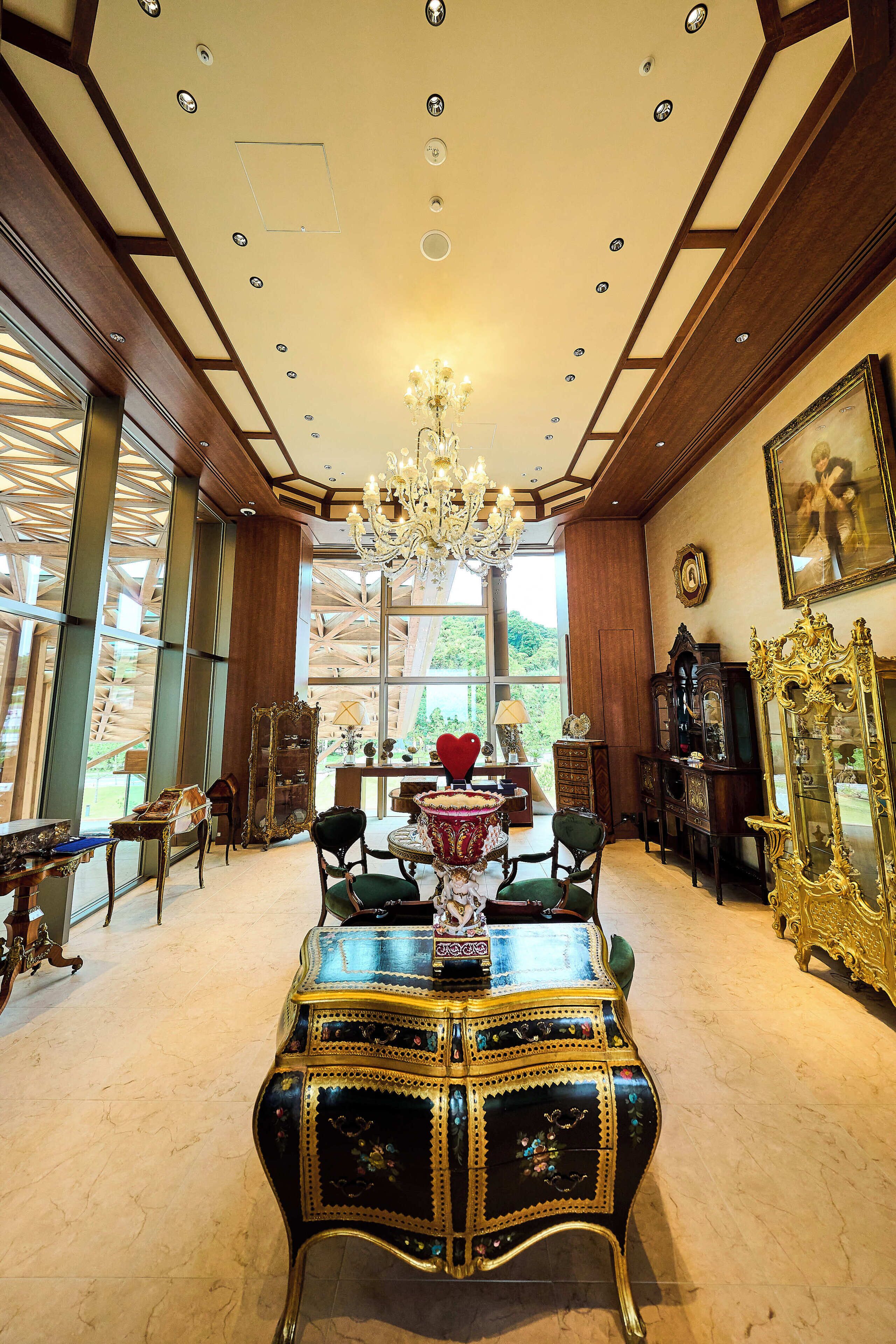 A spacious room with a chandelier, ornate furniture, and large windows offering a view of greenery.