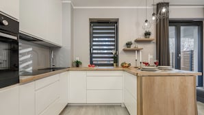 Μια μοντέρνα κουζίνα με λευκά ντουλάπια, έναν ξύλινο πάγκο, έναν εντοιχισμένο φούρνο και ένα παράθυρο με περσίδες.