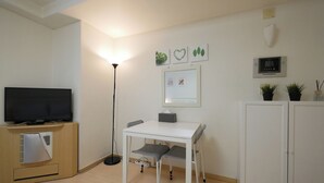 Une petite pièce bien éclairée, équipée d’une télévision, d’un bureau et d’une chaise. Des tableaux sont accrochés au mur, et une petite plante y est également posée.