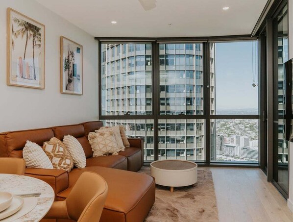 Ein modernes Wohnzimmer mit einem braunen Ledersofa, einem runden Couchtisch und einem großen Fenster mit Blick auf hohe Gebäude.