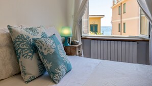A bed with patterned pillows, a nightstand with a lamp, and a view of buildings through a window. - PETITE PARAGGI - Sea View and Comfort (Paraggi)