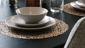 Una mesa de comedor dispuesta con platos, tazones y vasos blancos, un florero con plumas de plumas de pampa secas y una botella de perfume.