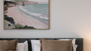 Una cama bien hecha con sábanas blancas y una manta marrón, cabecero con tres almohadas, y un cuadro de paisaje colgado encima de la cama.