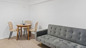 A modern, minimalist room with a grey sofa, two beige chairs, and a wooden dining table set for two. - Self-Contained Studio | Perfect for Solo Travelers (MacGregor)