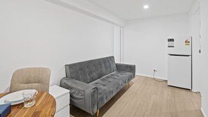 A modern, minimalistic room with a grey sofa, a small round table with a glass, and a white refrigerator. - Self-Contained Studio | Perfect for Solo Travelers (MacGregor)