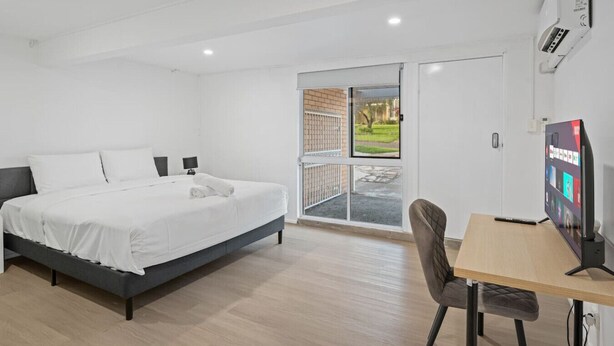 A modern bedroom with a bed, bedside tables, a desk with a chair, a TV, and a sliding glass door leading to an outdoor area. - Self-Contained Studio | Perfect for Solo Travelers (MacGregor)