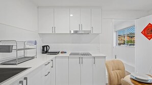 Een kleine keuken met witte kasten, een roestvrijstalen afzuigkap, een waterkoker en een eethoekje met een stoel en tafel.