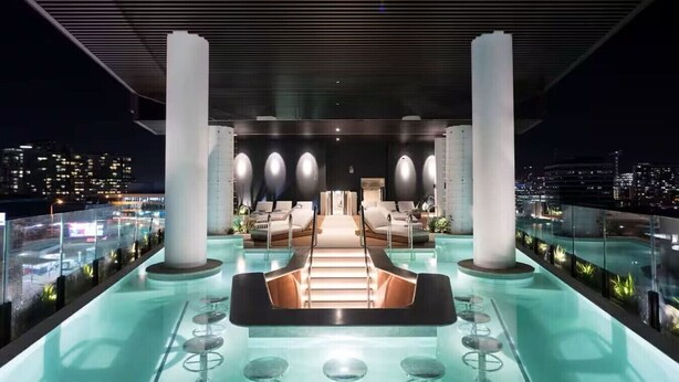 Un salón en la azotea con piscina, zonas de estar y vistas nocturnas a la ciudad.