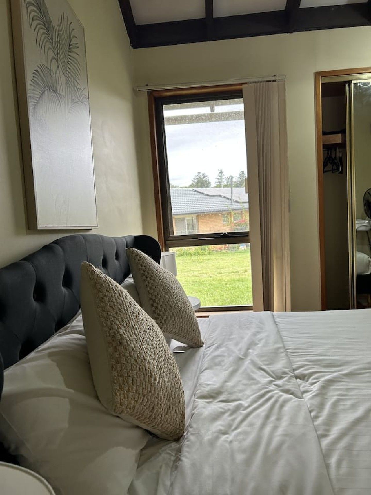 Ein Schlafzimmer mit Bett, einem Fenster mit Vorhängen, Blick auf ein Haus und einem Gemälde an der Wand.