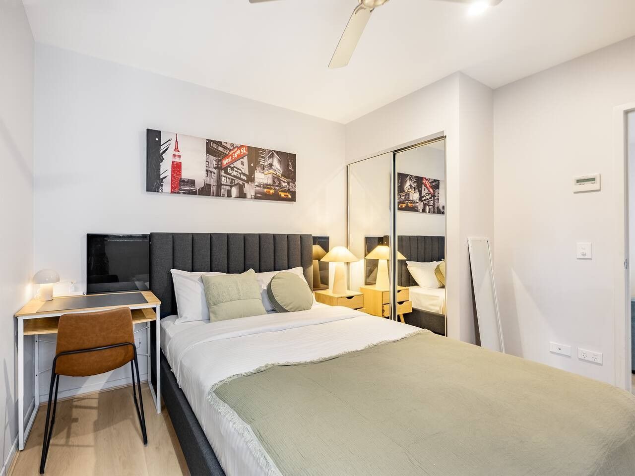 Habitació d'hotel moderna amb llit, escriptori, cadira, làmpades de nit i un mirall gran.