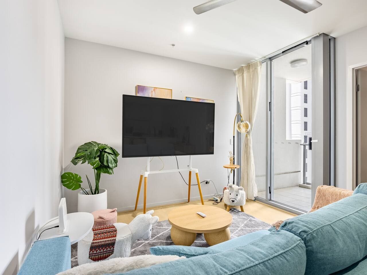 A modern living room with a flat-screen TV, a sofa, a coffee table, a potted plant, and a sliding glass door leading to an outdoor area.