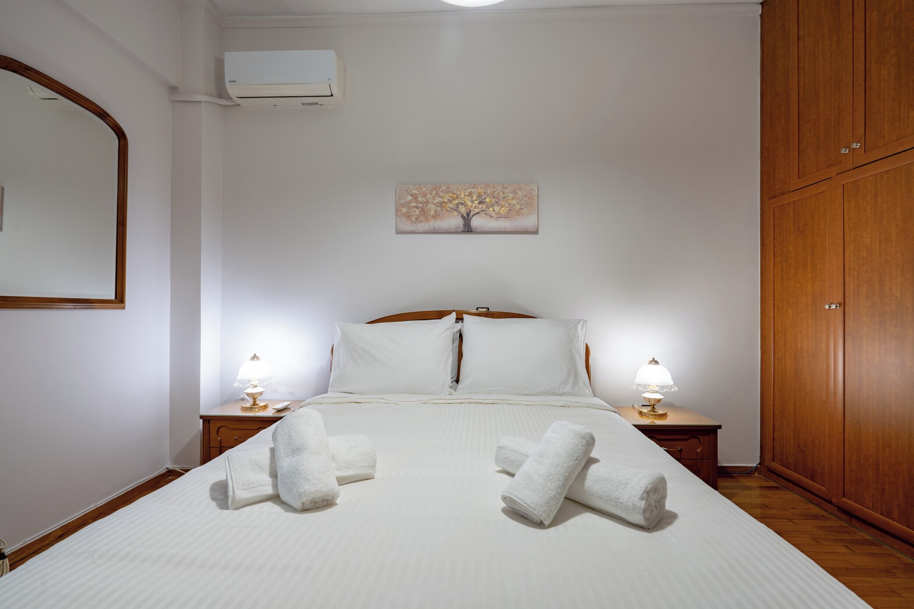 A hotel room with a bed, two bedside tables with lamps, a wooden wardrobe, a painting of a tree, and an air conditioning unit.