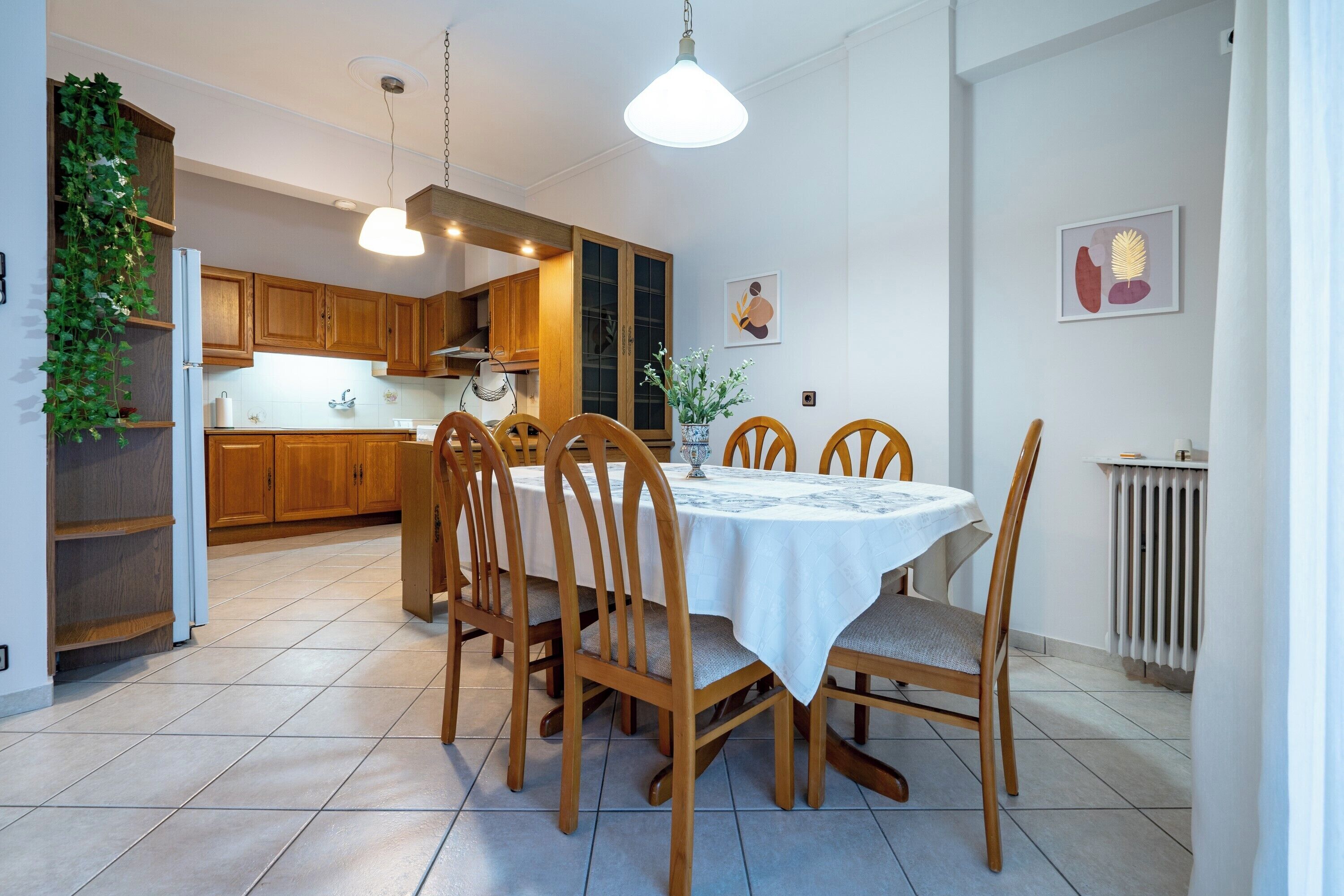 A dining area with a round table, wooden chairs, and a white tablecloth. There is a kitchen in the background with wooden cabinets and a hanging plant.
