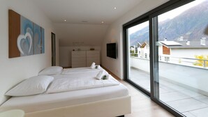 Une chambre moderne avec un grand lit, une commode et un téléviseur. Elle dispose d’un balcon avec vue sur les montagnes.