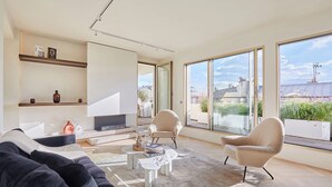 Een moderne woonkamer met een bank, twee fauteuils, een salontafel en een televisie. Grote ramen bieden uitzicht op de skyline van de stad.