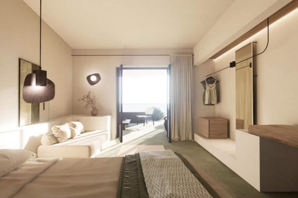 Ein modernes Hotelzimmer mit Bett, einer Sitzecke mit Sofa und Sesseln, einem kleinen Tisch, einem Spiegel und Blick ins Freie.