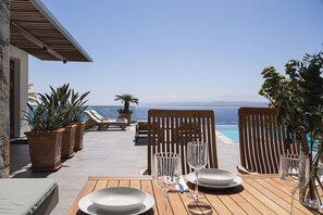 Ein Holztisch ist für eine Mahlzeit gedeckt, mit Blick auf das Meer und die dahinterliegenden Berge.