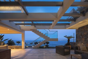 A modern interior with a staircase leading to a balcony overlooking the sea, furnished with wicker furniture and potted plants. - Candia Lux Villa with Secluded Beach by etouri (Heraklion)