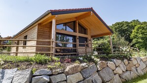 A wooden cabin with a sloped roof, large windows, and a stone wall in front. - Modernes Ebenerdiges Chalet im Vital Camp Bayerbach (Bayerbach)
