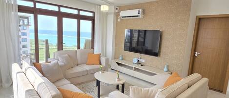 Una sala de estar moderna con un sofá modular, una mesa de centro, un televisor de pantalla plana montado en la pared y un ventanal con vistas al mar.