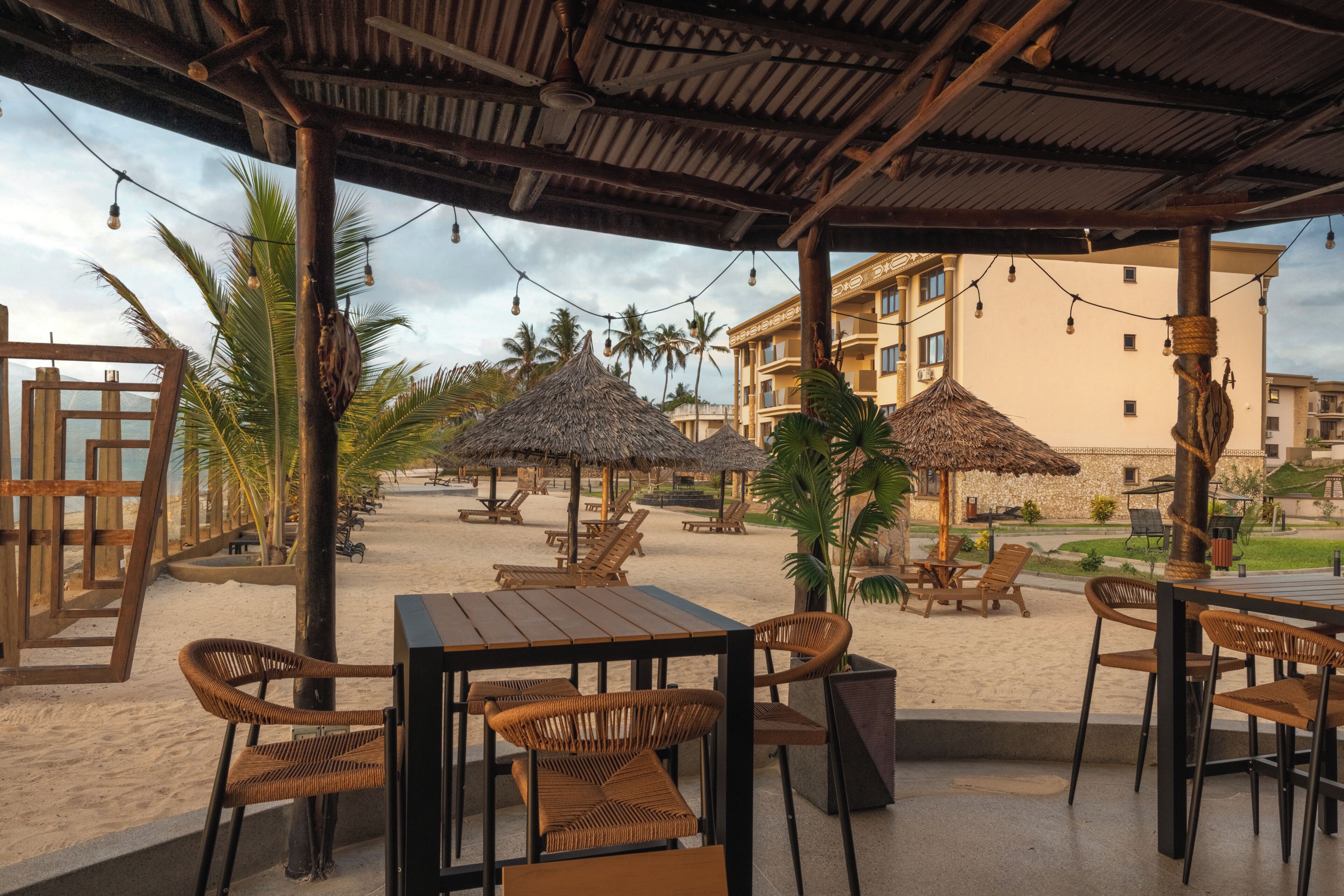 An outdoor seating area with tables and chairs, thatched umbrellas, and a view of a building in the background.