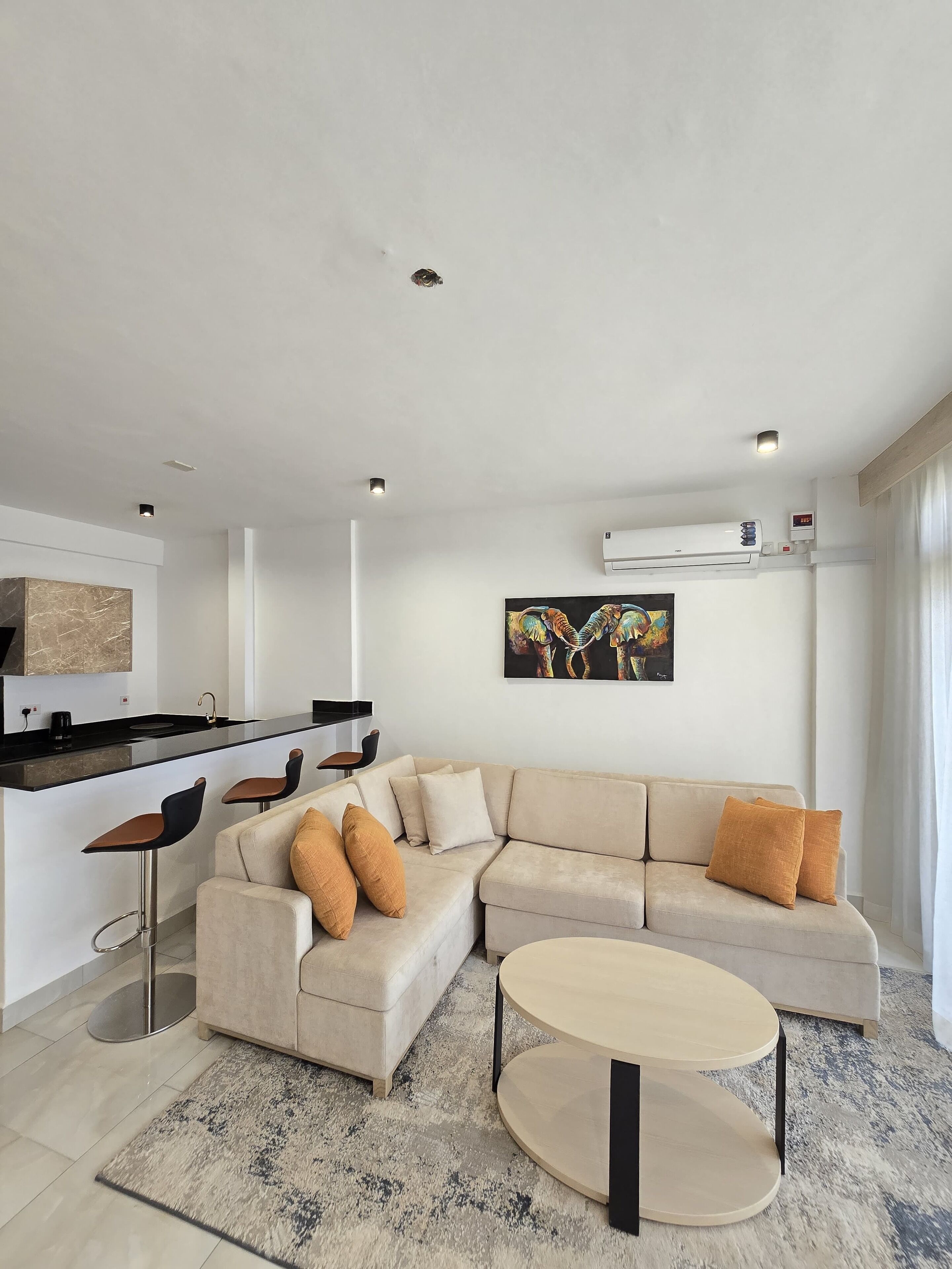 Una sala de estar moderna con un sofá seccional color beige, una mesa redonda y una cocina con encimera negra y taburetes de bar.