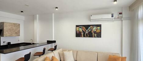 Una sala de estar moderna con un sofá seccional color beige, una mesa redonda y una cocina con encimera negra y taburetes de bar.