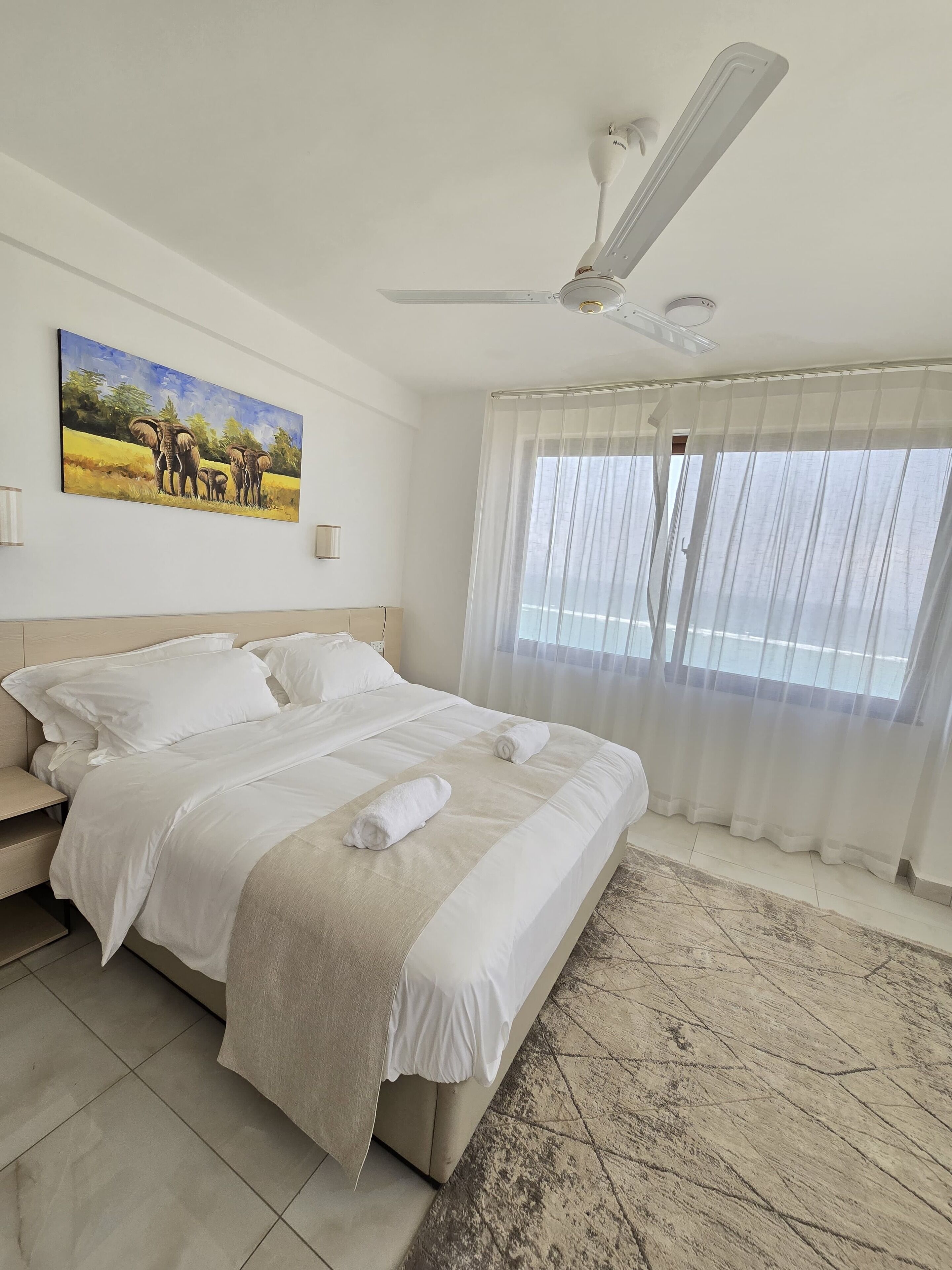A spacious bedroom with a large bed, a ceiling fan, a painting of elephants, and a view through sheer curtains.