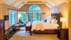Ein Schlafzimmer mit einem großen Bett, einem Sessel und einem Schreibtisch. Es gibt große Fenster und eine Holzbalkendecke.