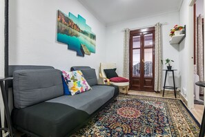 Un soggiorno con un divano grigio, cuscini colorati, un tappeto a motivi, un quadro appeso, una porta in legno e una piccola pianta su uno scaffale.