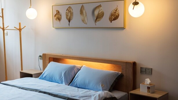 A neatly made bed with blue bedding, a wooden headboard, and a wall-mounted artwork of leaves.