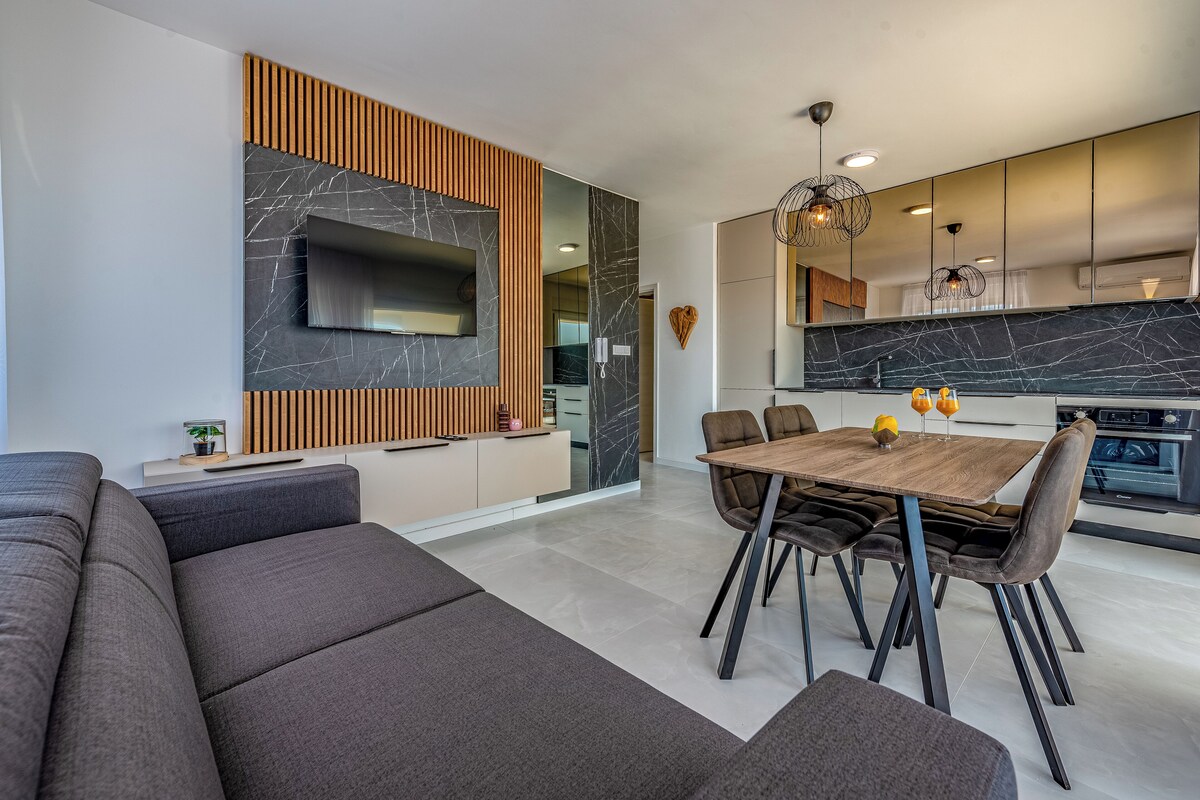 A modern living room with a large sectional sofa, a dining table with chairs, and a kitchen area with a large island and a built-in oven.