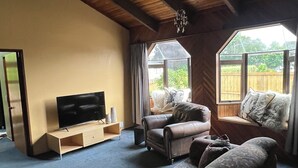 Un soggiorno con soffitto in legno, una televisione su un supporto in legno, un divano, una poltrona e una finestra vista su spazi verdi.