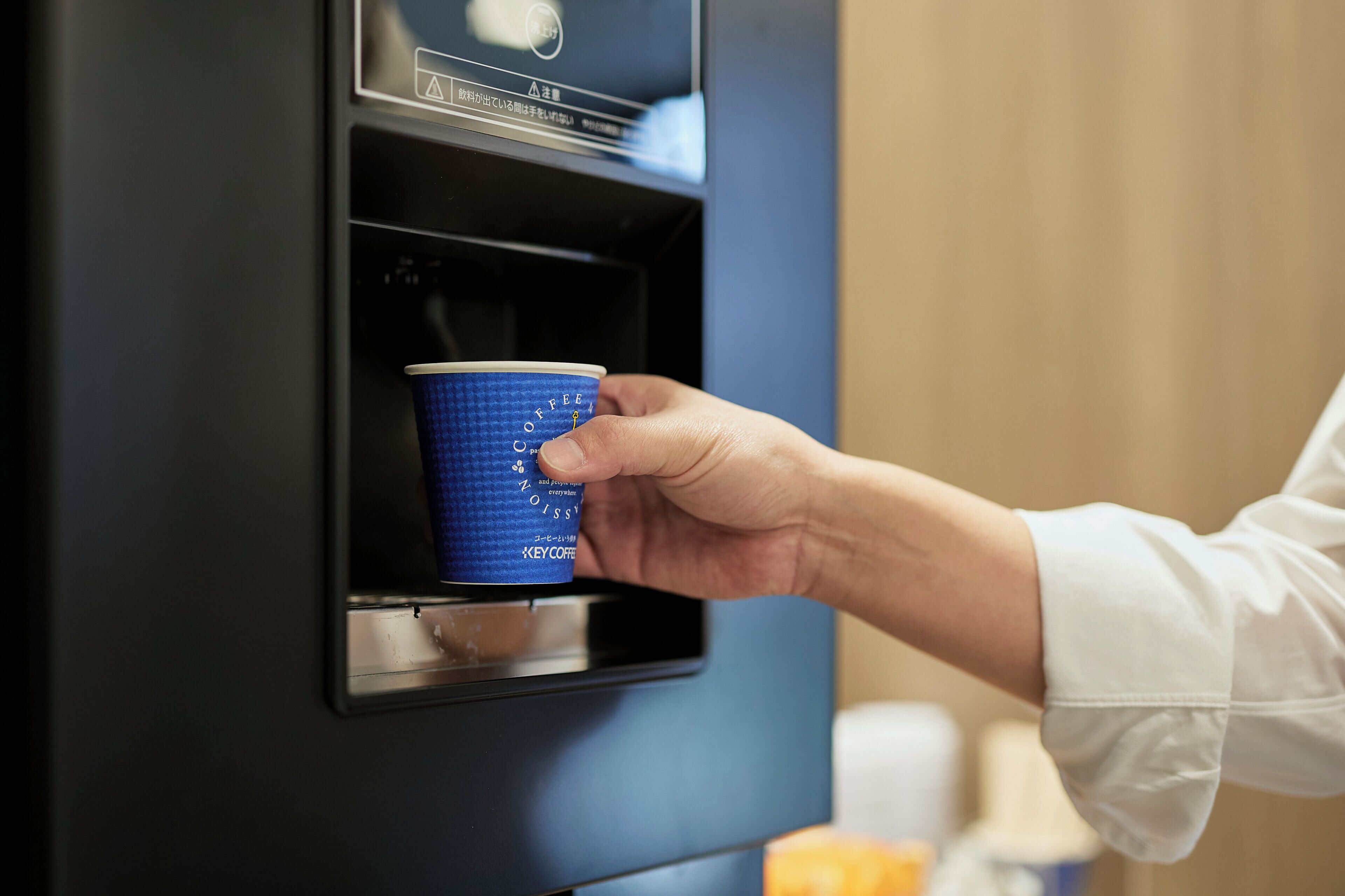 Mão segurando um copo descartável azul em frente a uma máquina de café.