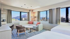 Quarto de hotel com sofá, mesa pequena, cadeira, cama e grandes janelas com vista para a cidade.