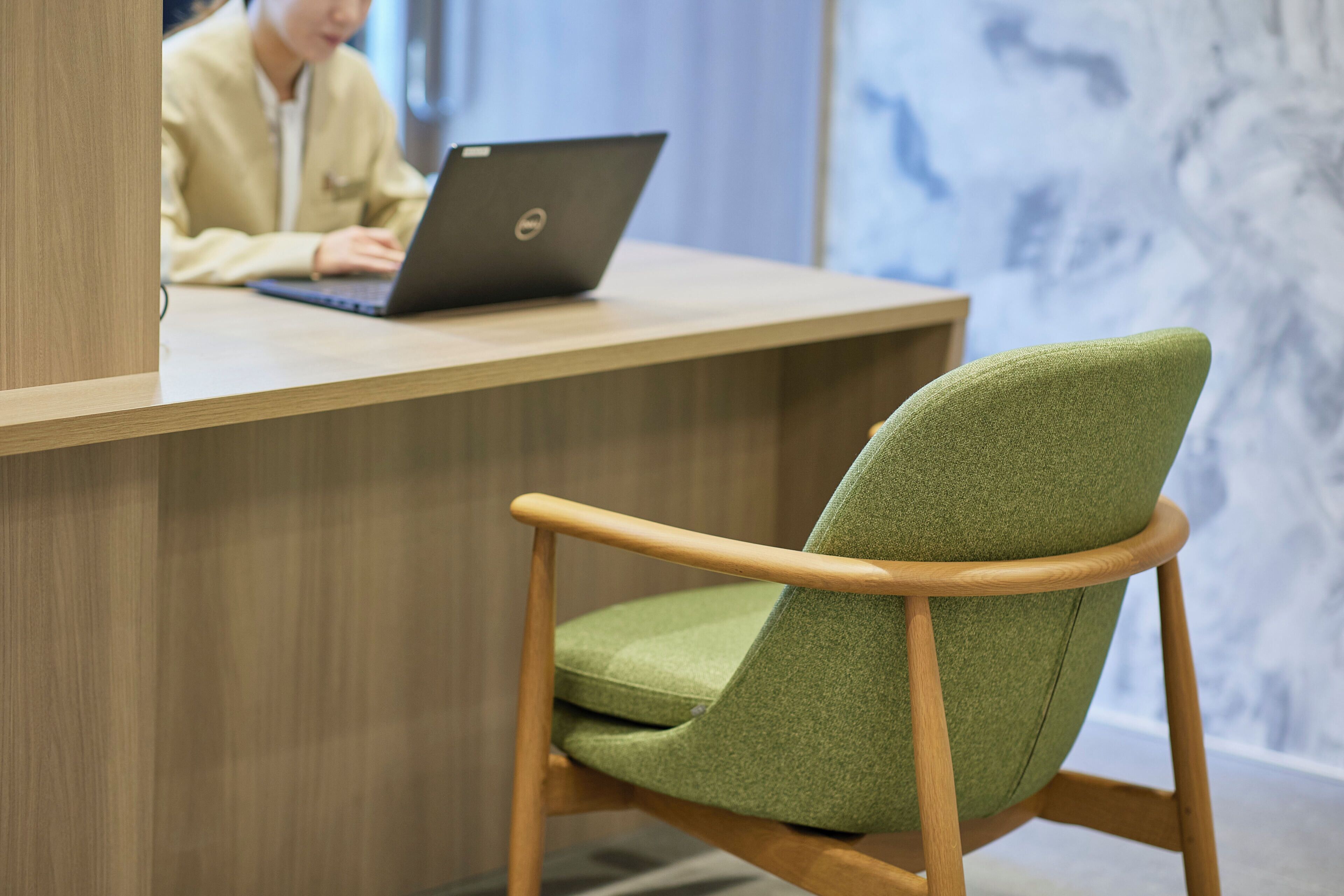 A wooden reception desk with a laptop, a green upholstered chair, and a marble wall in the background.