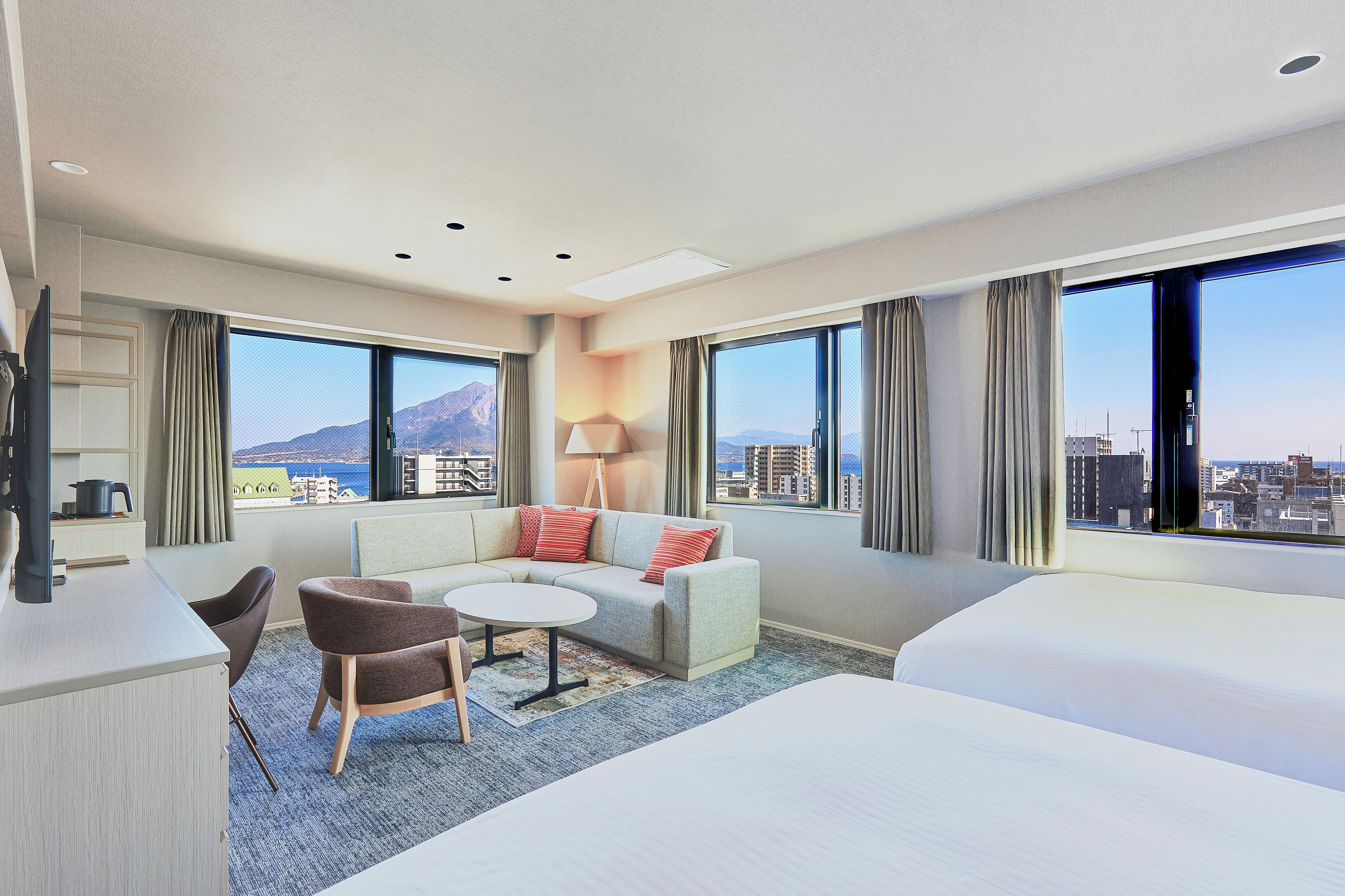 Habitación de hotel con cama, sofá, una mesita y una silla. Amplias ventanas con cortinas y vistas a edificios.