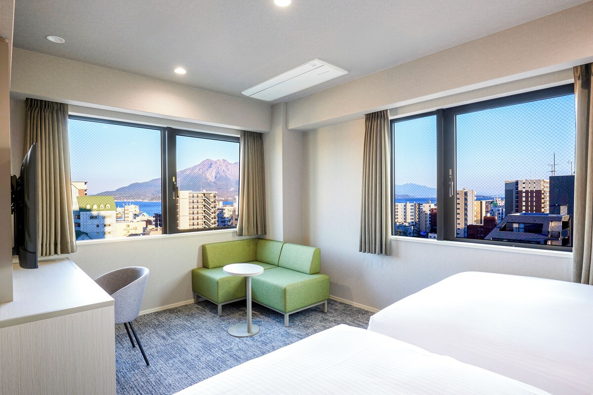 A hotel room with a bed, a green sofa, a small table, a chair, and large windows offering a city view.