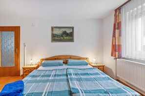 Ein Schlafzimmer mit Holz-Kopfteil, zwei Nachttischen mit Lampen, einem Bett mit blau-weißer Bettwäsche, einem gerahmten Landschaftsbild an der Wand und einem Fenster mit Vorhängen.
