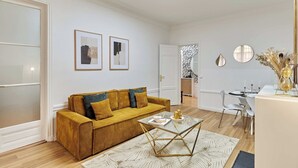 Un salon moderne avec un canapé de couleur moutarde, une table basse en verre et un coin repas avec des chaises blanches.